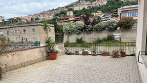 A rooftop terrace with potted plants, a tiled floor, and a view of a hillside with buildings.