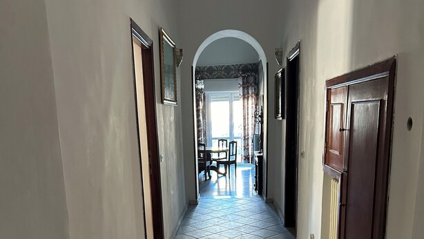 A hallway with tiled flooring, a dining area with chairs and a table, and a large arched doorway leading to another room.