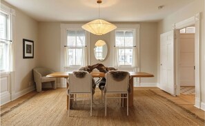 A dining room with a wooden table, four chairs with fur trim, a hanging light fixture, and large windows with white curtains. - 3-bedroom Gem on Main Street Sag Harbor Village (Sag Harbor)