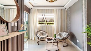 A modern living room with a round mirror, two cushioned chairs, a coffee table, and a large window with curtains.
