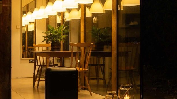 Una moderna hall d'hotel con parete in vetro, diverse lampade a sospensione, un tavolo in legno, sedie e una valigia.