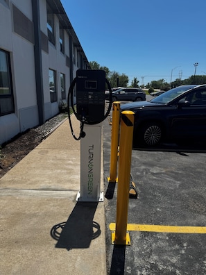 Uma área de estacionamento com uma estação de carregamento identificada como 'TURNING GREEN' e um edifício ao fundo.