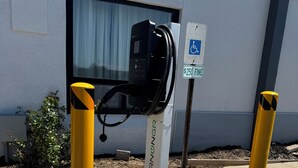 Un parquímetro con un letrero de accesibilidad para sillas de ruedas, bolardos amarillos y un edificio con una ventana al fondo.