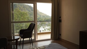 A hotel room with a bed, a chair, a table, and a large sliding glass door leading to a balcony with a view of a forested hillside. - Ramana on the Ganges (Tapovan, Rishikesh)