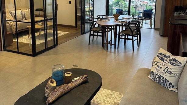 Un soggiorno moderno con divano, tavolino, zona pranzo e vista sulla città attraverso ampie finestre.