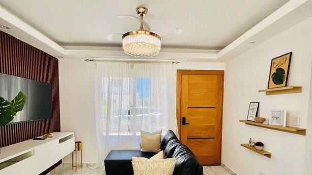 A modern living room with a black leather sofa, a TV mounted on the wall, a chandelier, and a wooden door. - Private swimming pool in punta cana, electricity included (Punta Cana)