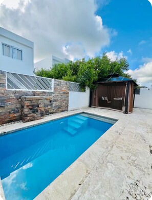 Ein Pool mit Steinmauern, eine überdachte Sitzgelegenheit und ein modernes Gebäude mit großen Fenstern.