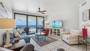 A living room with a sofa, armchair, coffee table, TV, and a view of the ocean. - Summer House Condo 503B (Orange Beach)