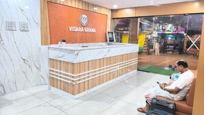 Una recepción con encimera de mármol y panel de madera, un hombre sentado en un sofá con una maleta, y una tienda visible a través del muro de cristal.