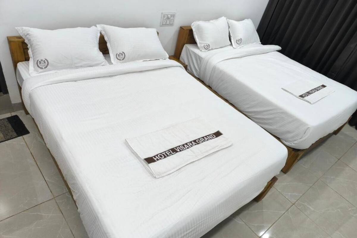 Dos camas individuales con ropa de cama y almohadas blancas, un cabecero de madera, una mesita de noche pequeña y una toalla con la marca 'HOTEL VISARA GRAND'.