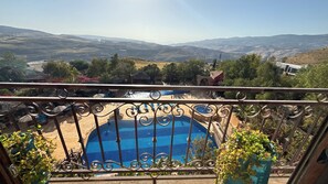 A balcony with a view of a swimming pool, surrounded by greenery and distant hills.