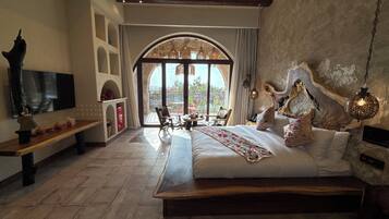 A bedroom with a large bed, a TV, a seating area with chairs, and a view through a large arched window.