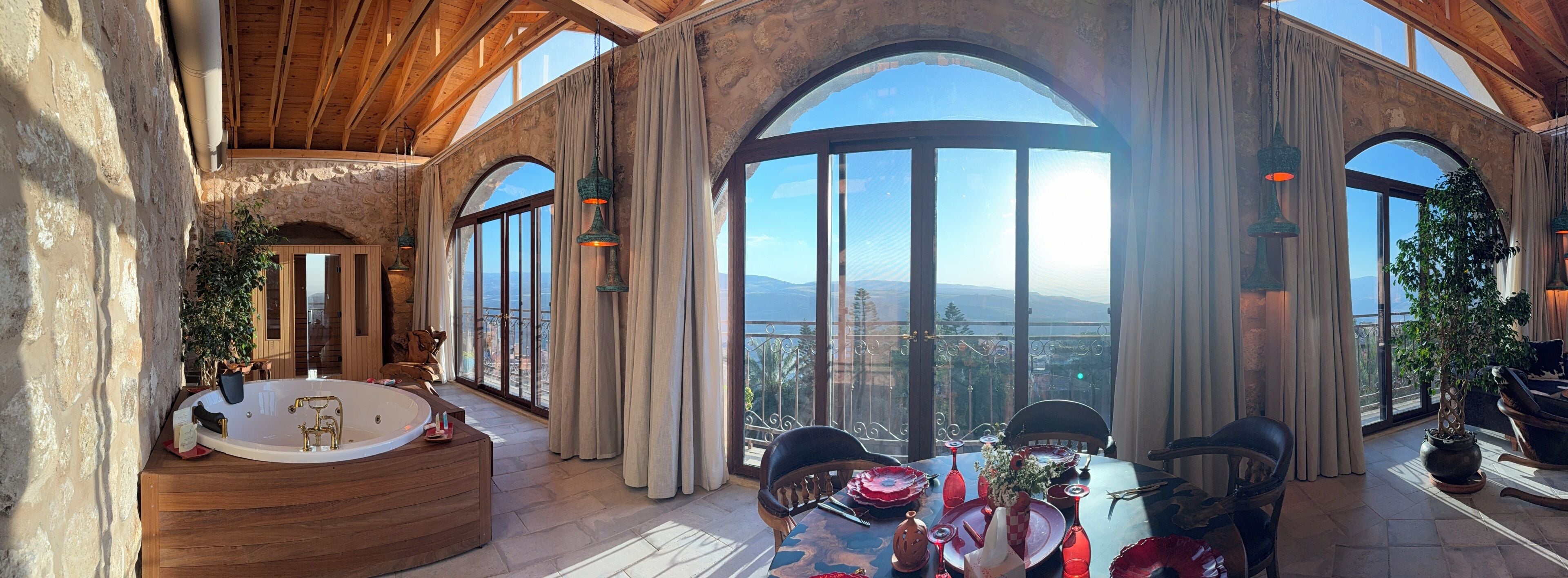 A spacious room with a large bathtub, a dining table set for two, and a view of the mountains through large windows.