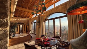 A spacious room with a high ceiling, large windows, a wooden table set for dining, and a stone wall.