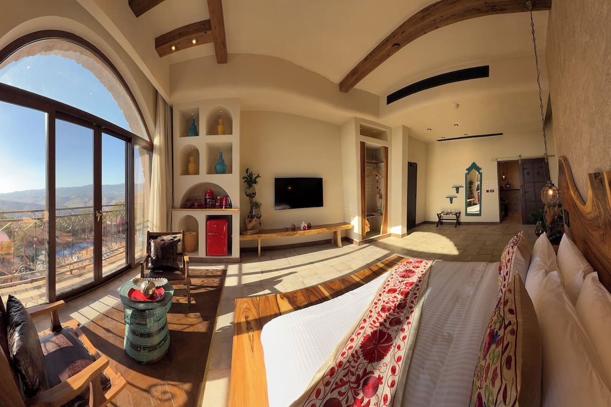 Una habitación amplia con un ventanal grande, una cama, un televisor y techo de madera.