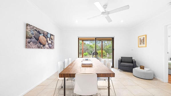 A modern dining room with a wooden table, white chairs, a ceiling fan, and a view through glass doors to an outdoor area.