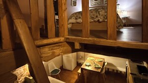 Un intérieur rustique avec un escalier en bois, un petit coin repas avec une table dressée pour deux personnes, et un lit confortable doté d’une tête de lit à motifs.