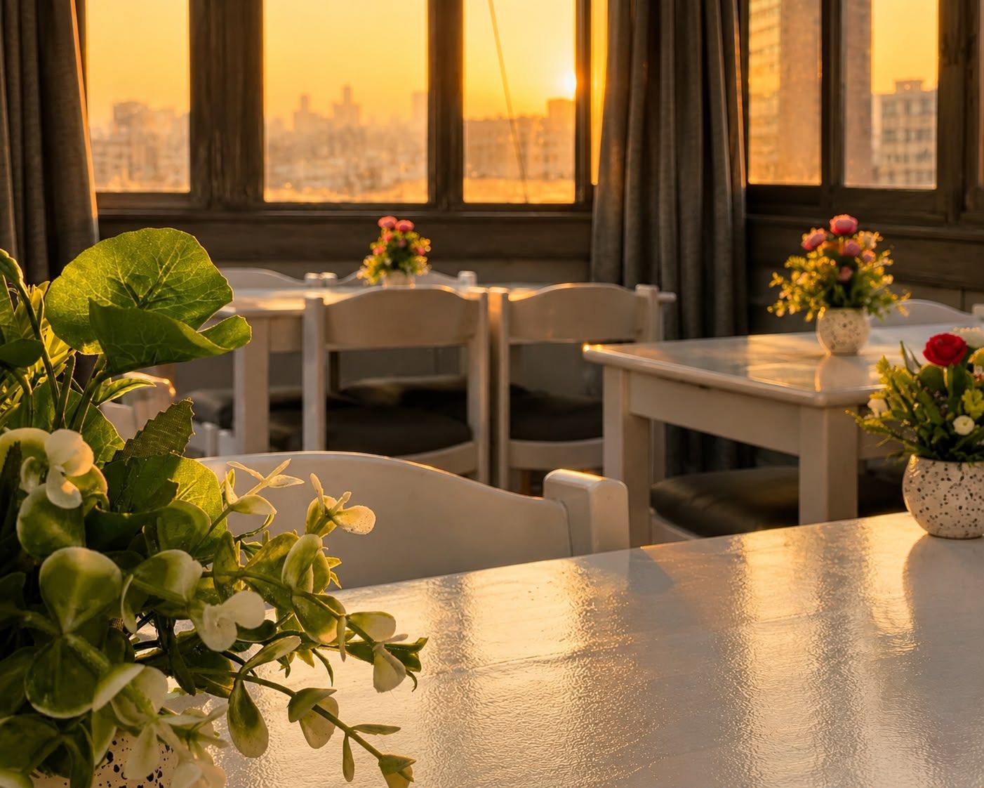 A dining area with tables and chairs, a city view through large windows, and potted plants on the tables.