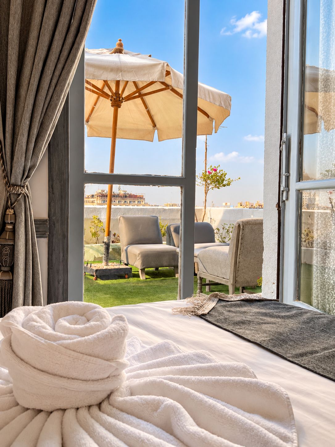 Ein Zimmer mit großem Fenster, einem Bett mit zusammengerolltem Handtuch und Blick auf die Stadtlandschaft.