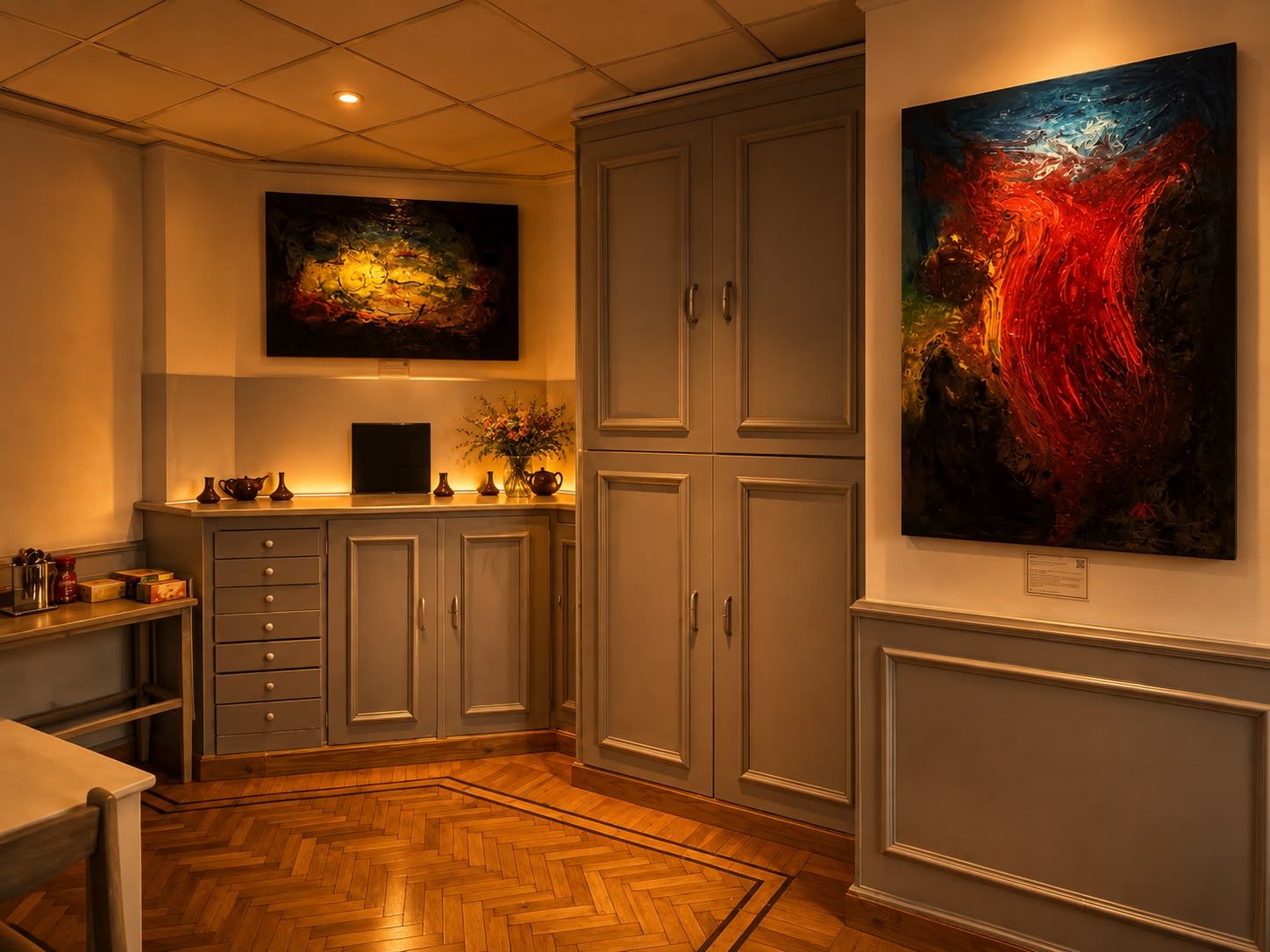 A kitchen with a herringbone floor, grey cabinets, and two paintings on the walls.