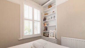 A bedroom with a bed, a window with shutters, a bookshelf, and a radiator. - Spacious East London Home with a beautiful Garden - Pass the Keys (London)