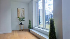 Ein Zimmer mit einem großen Fenster, einem Blumentopf, einem kleinen Tisch und einem Gemälde an der Wand.