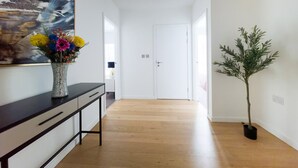 Un pasillo con piso de madera, una puerta blanca al final, una consola negra con cajones y un jarrón con flores de colores.