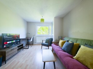 Ein Wohnzimmer mit Fernseher, Schreibtisch, Stuhl, Sofa und Fenster.