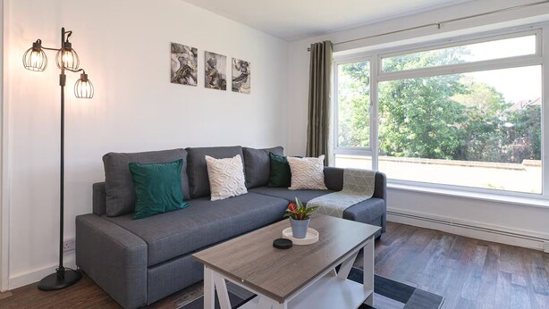 Ein modernes Wohnzimmer mit grauem Sofa, Couchtisch und Stehlampe. An der Wand hängen drei abstrakte Gemälde und ein großes Fenster mit Blick auf Grünflächen.