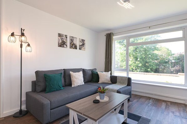 Ein modernes Wohnzimmer mit grauem Sofa, Couchtisch und Stehlampe. An der Wand hängen drei abstrakte Gemälde und ein großes Fenster mit Blick auf Grünflächen.