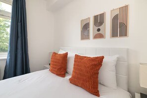 Ein Hotelzimmer mit einem weißen Bett, zwei orangefarbenen Kissen, einem weißen Kopfteil und drei abstrakten Wandbildern.