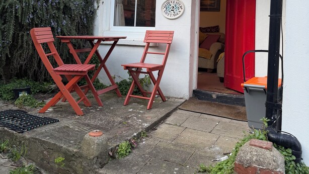 Un bâtiment blanc avec une porte rouge, un petit jardin avec une table et des chaises, et un mur en briques.