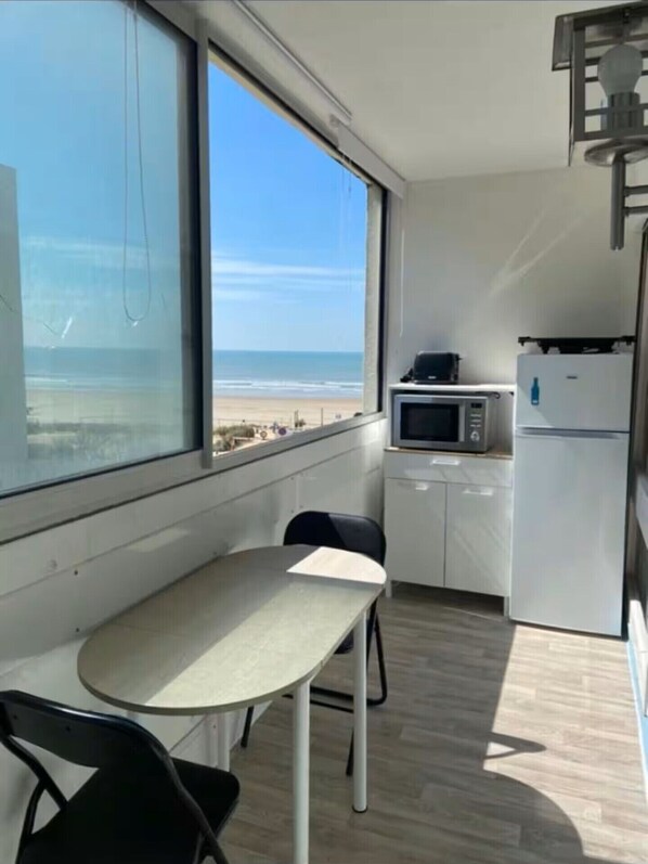 Eine kleine Küche mit Mikrowelle, Kühlschrank und einem Tisch mit Stühlen. Ein großes Fenster bietet Blick auf den Strand.