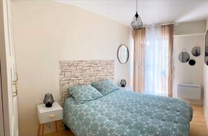 Ein Schlafzimmer mit einem Kopfende aus Steinoptik, einem Bett mit floralem Überwurf, Nachttischlampen, einem Nachttisch, einem Spiegel und einem Heizkörper.