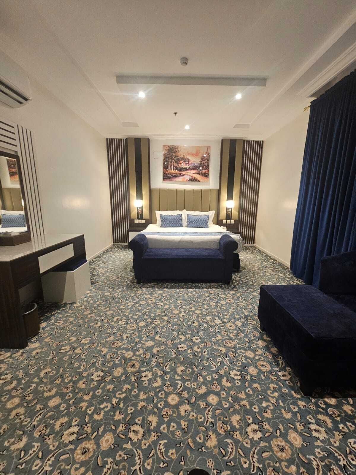 A hotel room with a bed, bedside tables, a desk, a chair, a patterned carpet, and a painting on the wall.