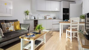A modern kitchen with white cabinets, a dining area with a wooden table and chairs, a sofa with patterned cushions, and a small coffee table. - Liner Apartment 17 (Falmouth)