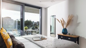A modern bedroom with a large bed, a bedside table with a vase, and a view of the outdoors through a sliding door. - Liner Apartment 17 (Falmouth)