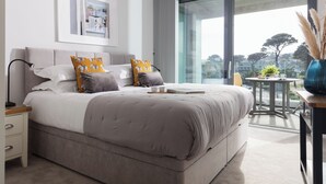 A modern bedroom with a large bed, a bedside table, a small round table with a plant, and a view of the outdoors through a sliding door. - Liner Apartment 17 (Falmouth)