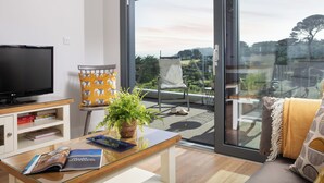 A modern living room with a flat-screen TV, a glass coffee table, a sofa, and a large sliding glass door leading to an outdoor balcony with a view of the landscape. - Liner Apartment 17 (Falmouth)