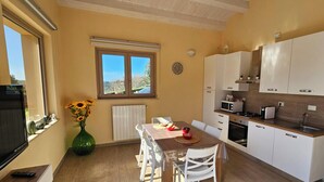 Cozinha compacta com armários brancos, uma mesa de jantar com cadeiras, uma janela grande com vista e um vaso com girassóis.