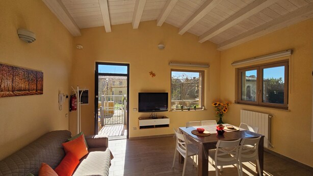 Uma sala de estar com sofá, televisão, mesa de jantar com cadeiras e janela com vista para o exterior.