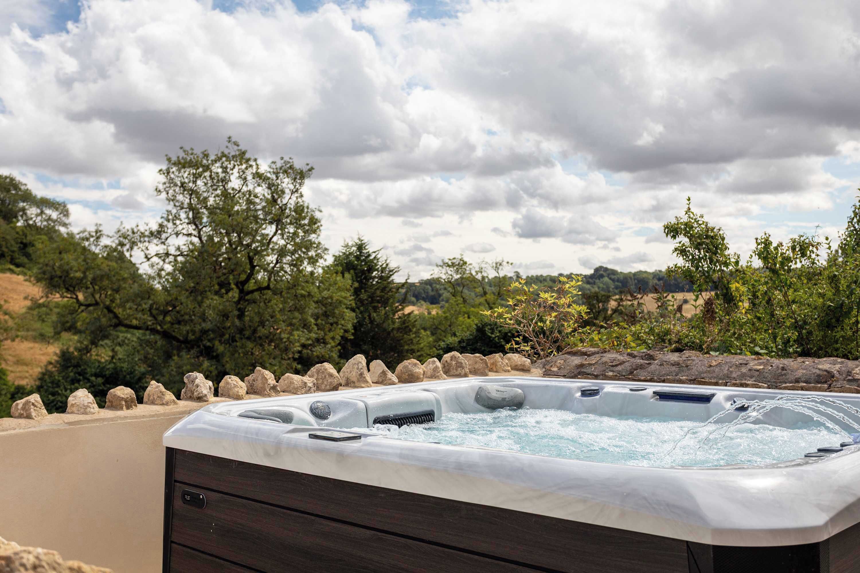 A hot tub with a wooden cabinet, surrounded by rocks and trees, set against a cloudy sky.