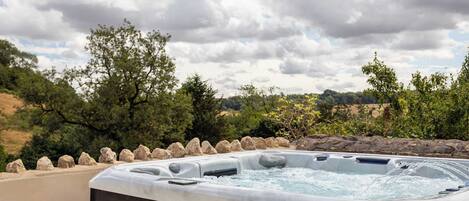 A hot tub with a wooden cabinet, surrounded by rocks and trees, set against a cloudy sky.