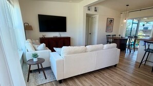 Ein geräumiges Wohnzimmer mit einem großen weißen Sofa, einem Flachbildfernseher, Holzböden und einem Essbereich mit Tisch und Stühlen.
