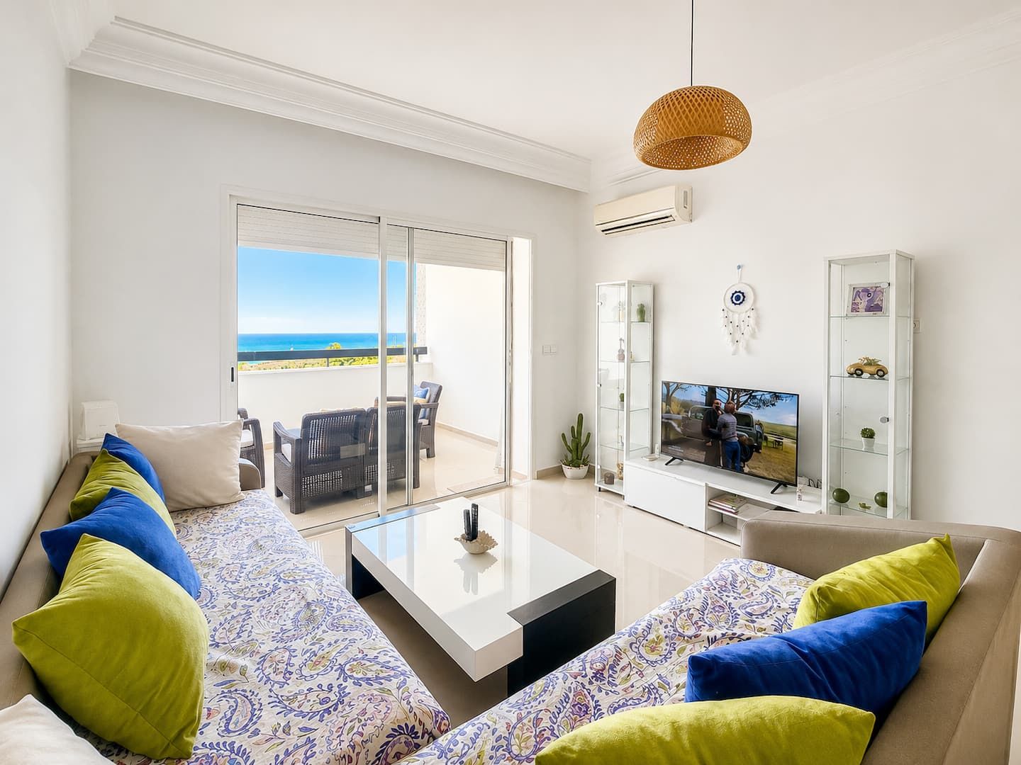 Un salon avec un canapé, une table basse et un téléviseur, un balcon avec vue sur l’océan et un attrape-rêves accroché au mur.