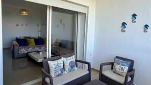 A room with a sliding glass door leading to a balcony, wicker furniture, and decorative wall art. - Magnificent sea & golf views. Pools, Terraces (Asilah)