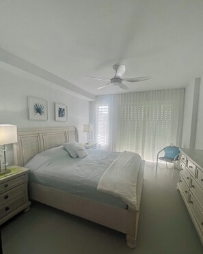 Ein Schlafzimmer mit Bett, Nachttisch, Kommode, Stuhl, Deckenventilator und einem Fenster mit Jalousien.