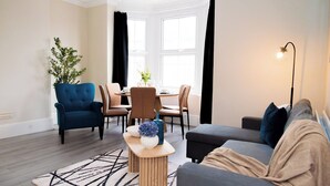 Een moderne woonkamer met een blauwe fauteuil, een grijze bank, een houten salontafel, en een eethoek met tafel en stoelen.
