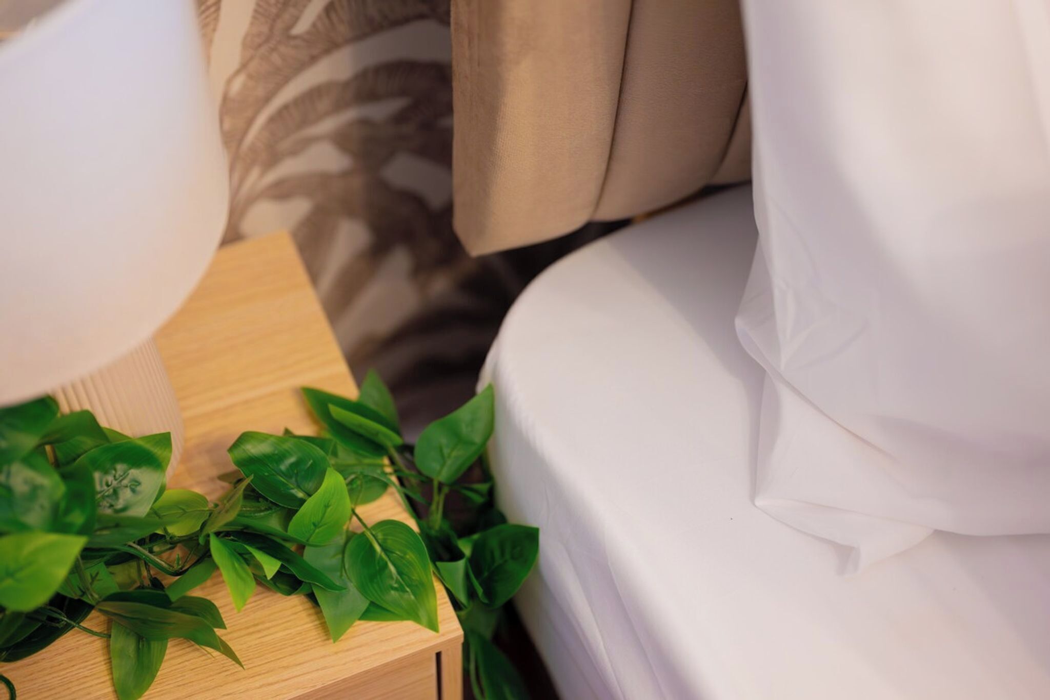 A bed with white bedding, a wooden nightstand with a plant, and a patterned pillow.