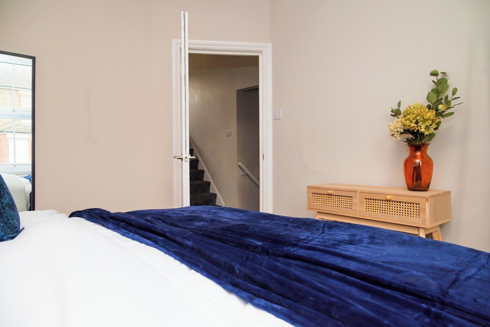 A bedroom with a bed, a wooden nightstand, a vase with flowers, and a door leading to a staircase.